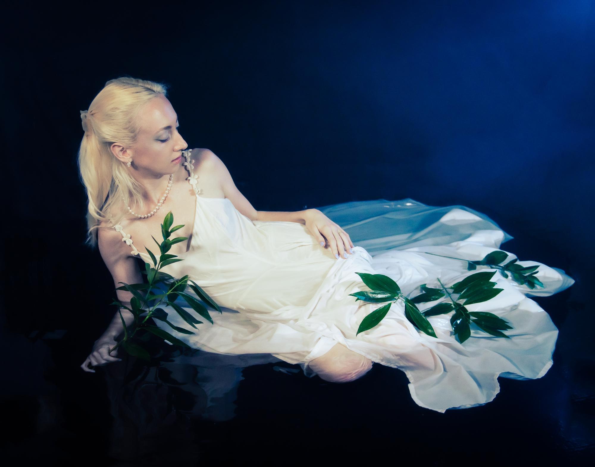 Female model sitting in pool of water with long white dress and leaves flowing out over her legs