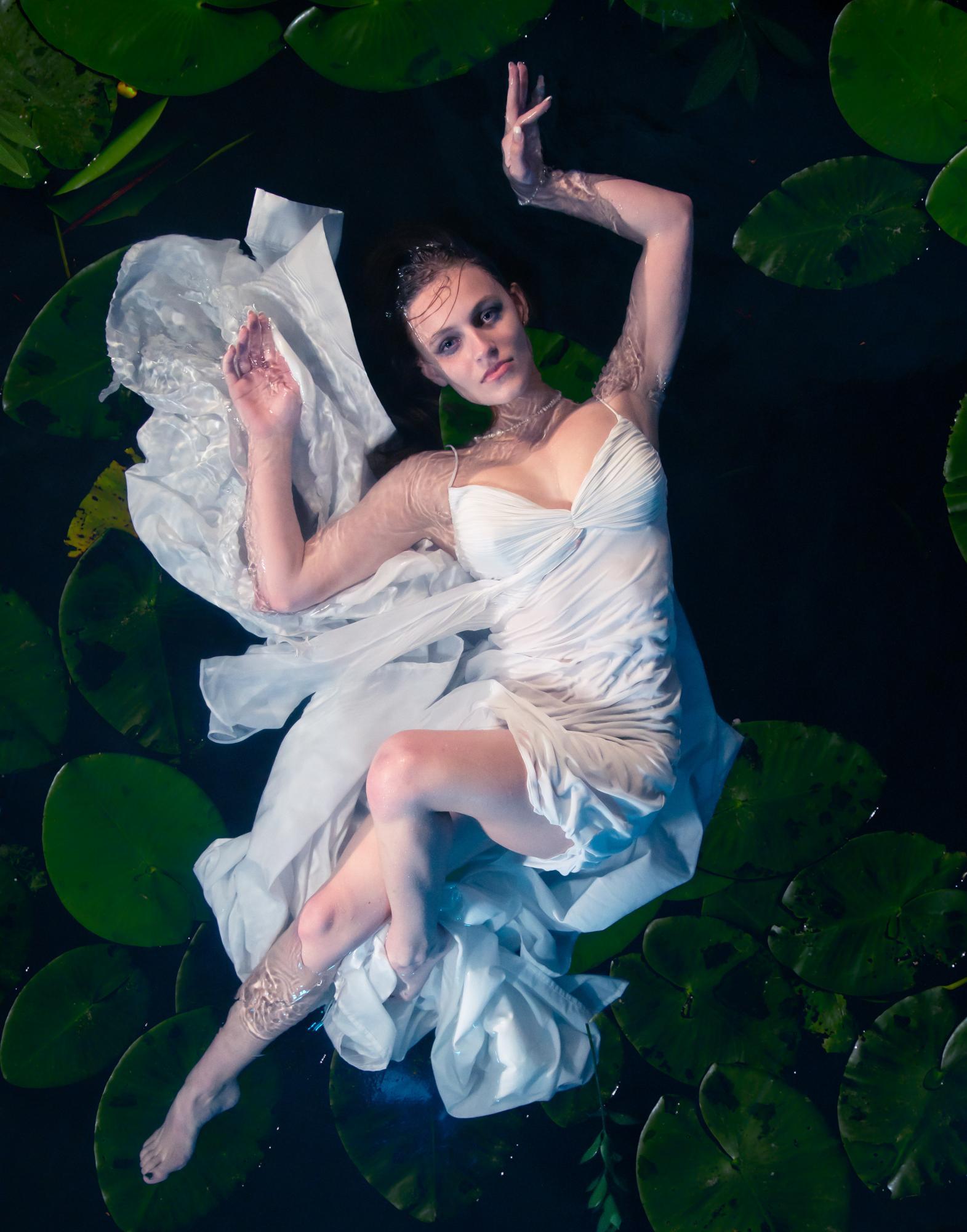 Picture taken from above of female model fully lying down in water with lily pads surrounding her in a flowing white dress