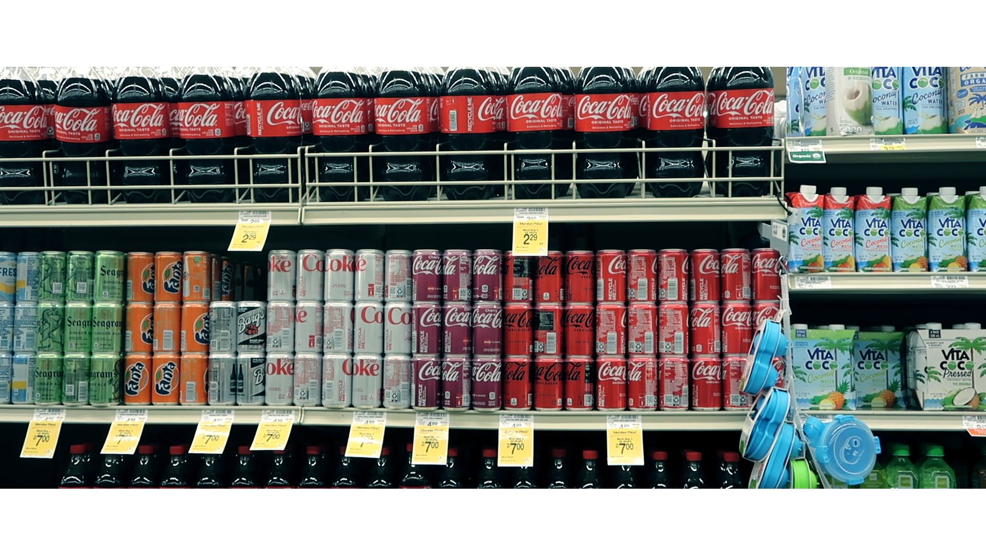 The beverage aisle of a grocery store. Several colors (red, white, orange, green) of cans and bottles of sodas are lined up throughout the frame.