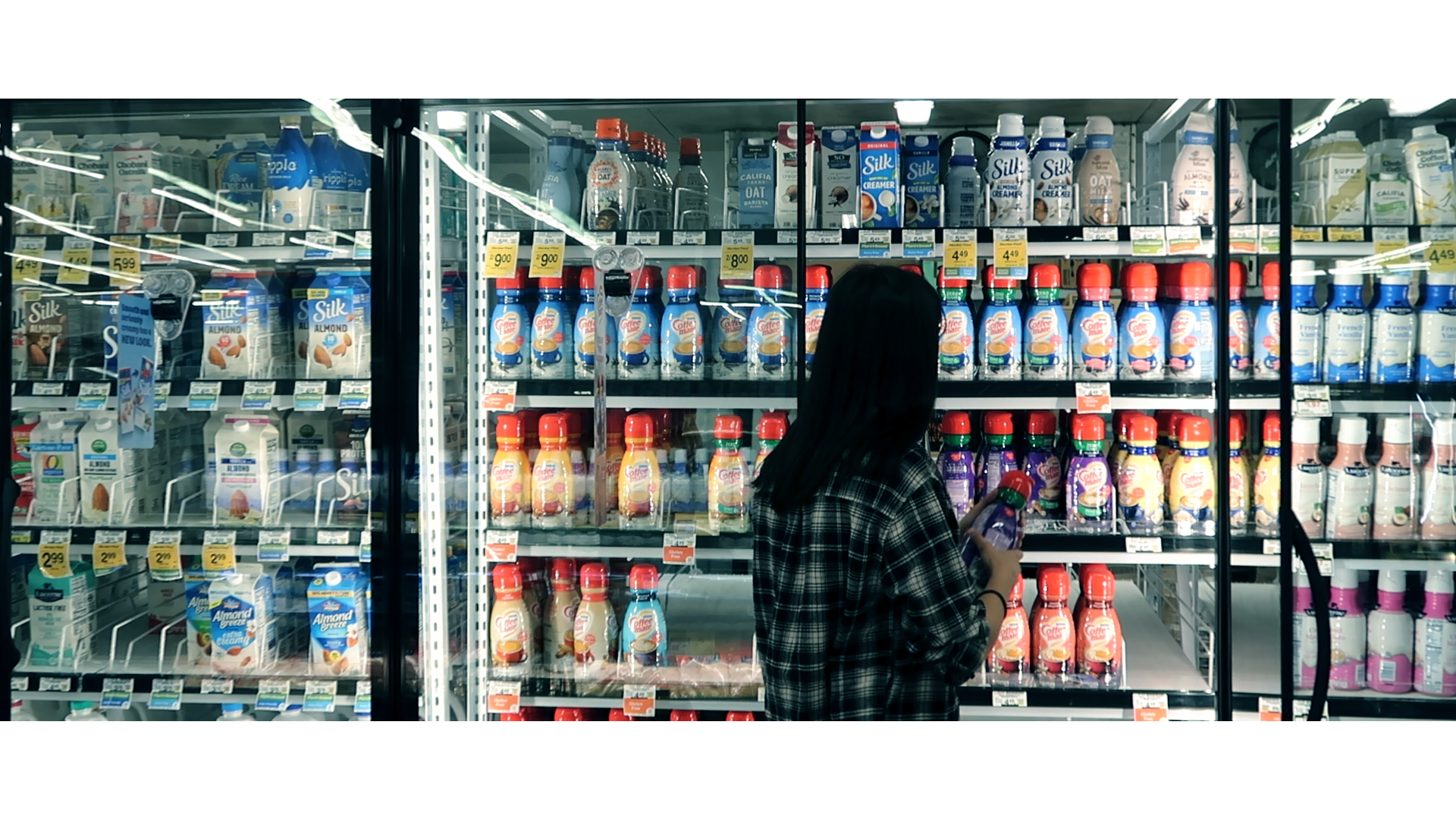 The milk/coffee creamer section of a grocery store. A girl in a black and white shirt is opening the fridge and is looking at the coffee creamer. The creamers are varying in vibrant, rainbow colors.
