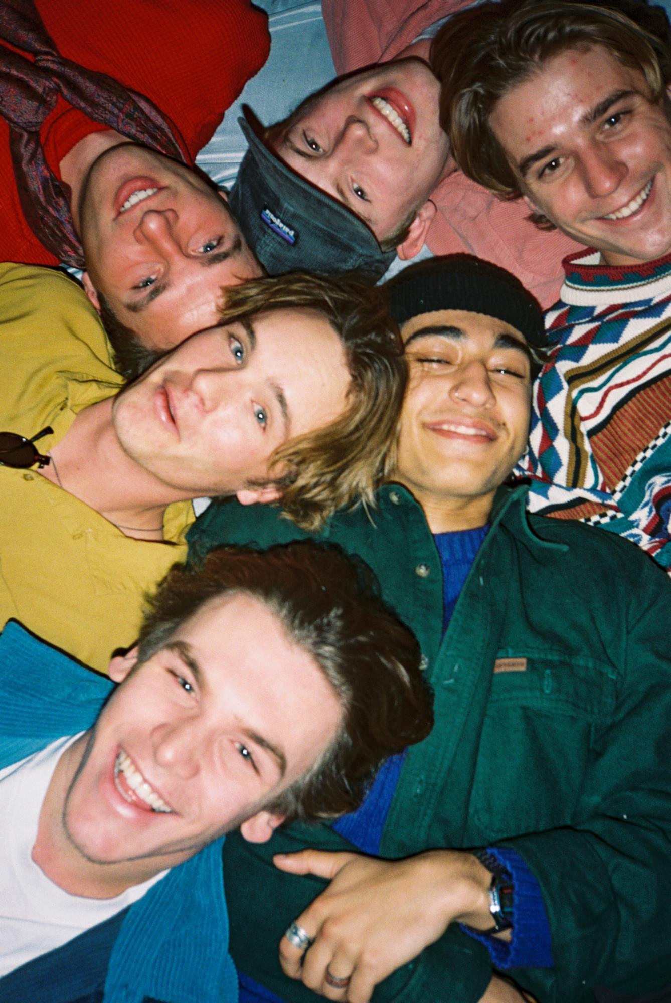 Just 6 six guys wearing their favorite outfit laying on the ground together smiling and looking up at a bird's eye position of the camera