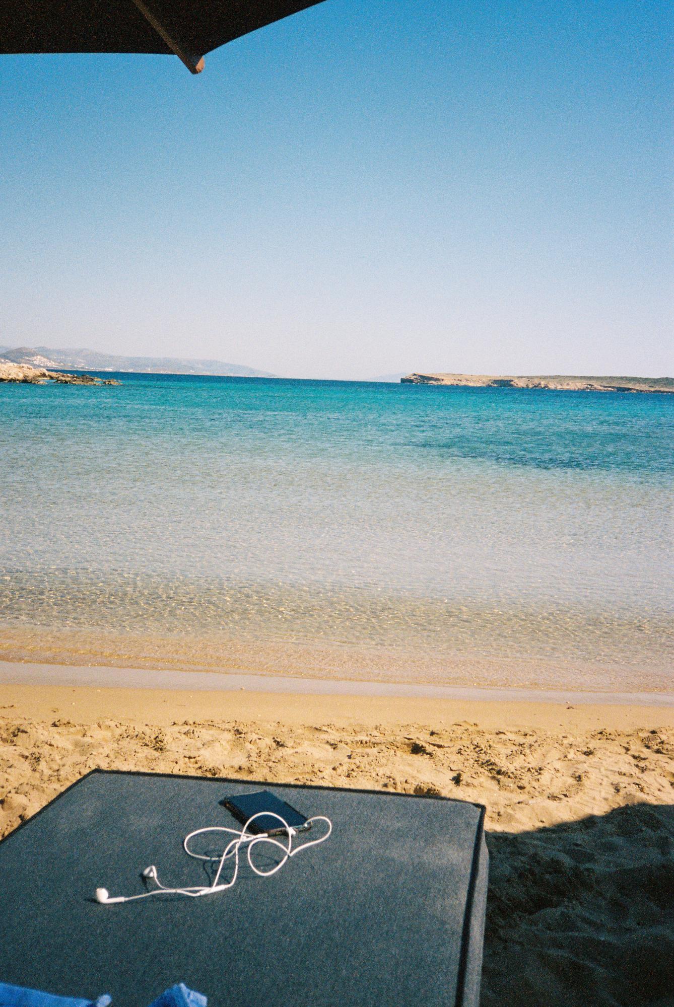 The calm, quiet beach in Paros Greece. The water is crystal clear turquoise, with a beach chair right up on the shore with an umbrella shading it