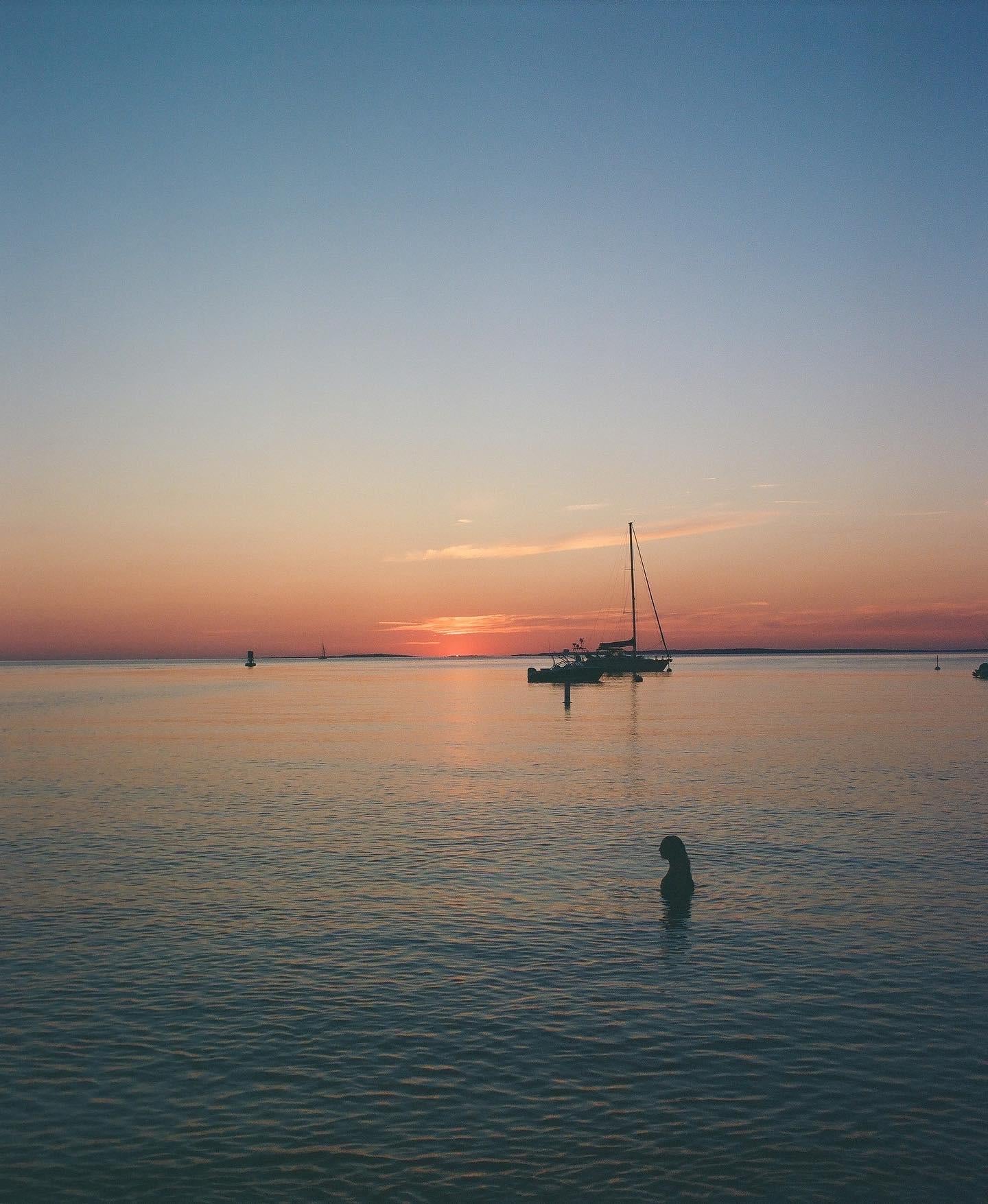 A silhouetted girl looks out across the calm seas as the sun just ducks beneath the horizon