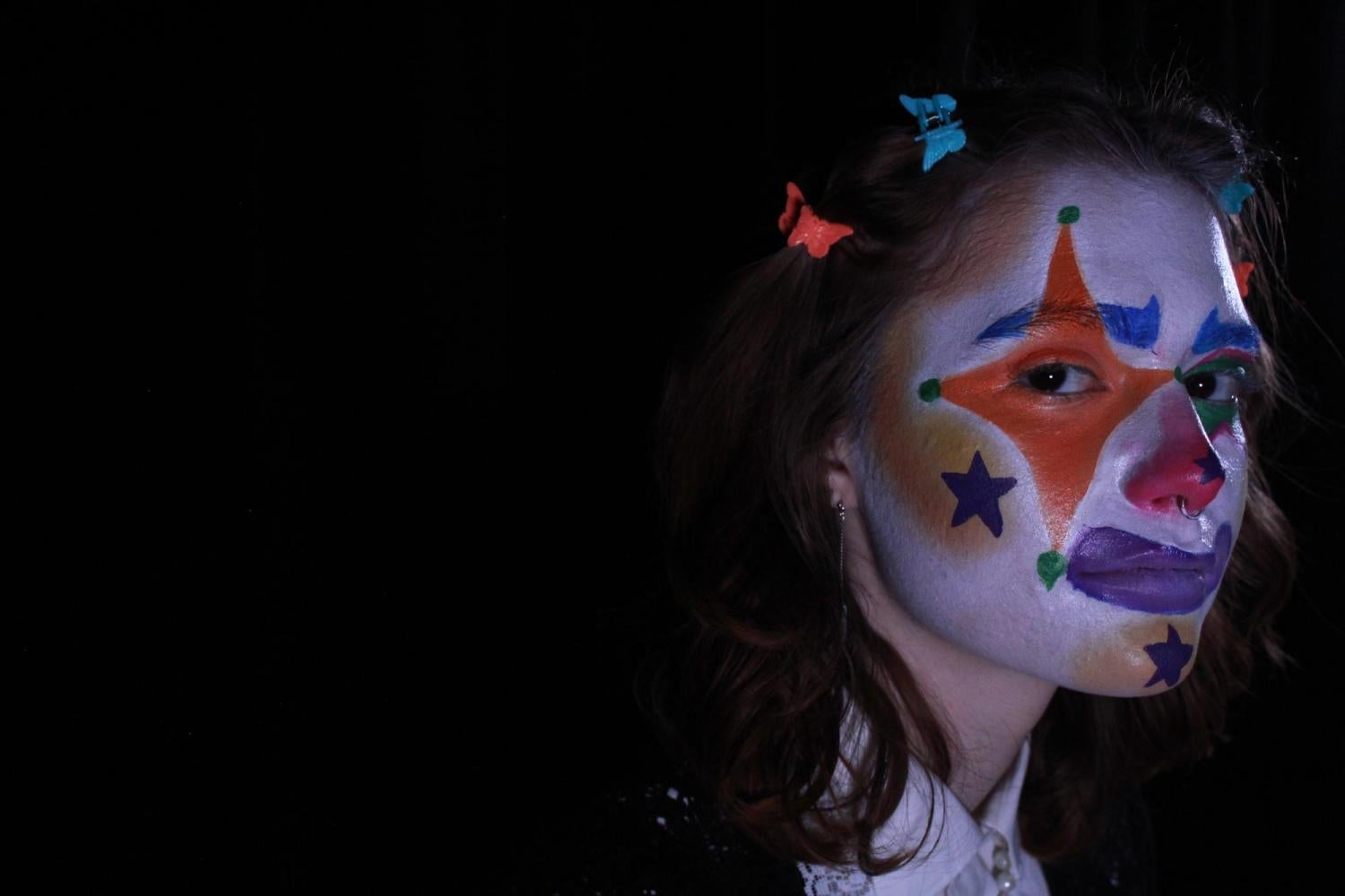 detailed makeup shot of orange star eye, purple smile, blue star on cheek, yellow cheek, blue eyebrows half turning to look at camera.