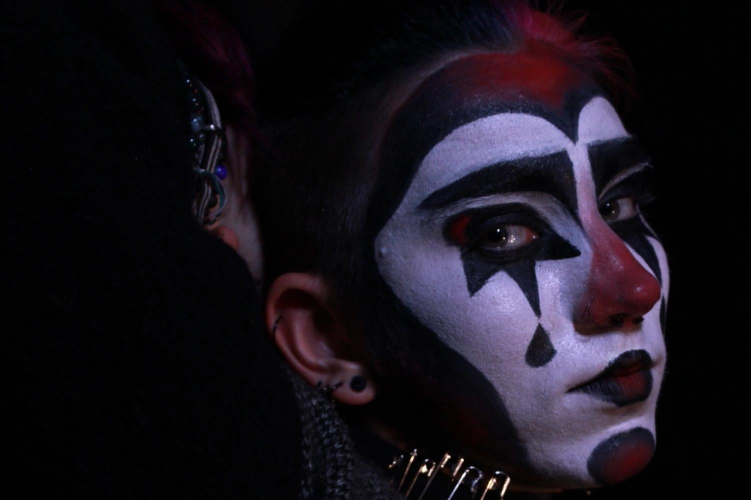 red and black makeup with teardrop on cheek laying on back, turning head to look at camera, arms folded behind head. sweater lying open, fish and "fuck" skeletal middle finger pins visible on sweater
