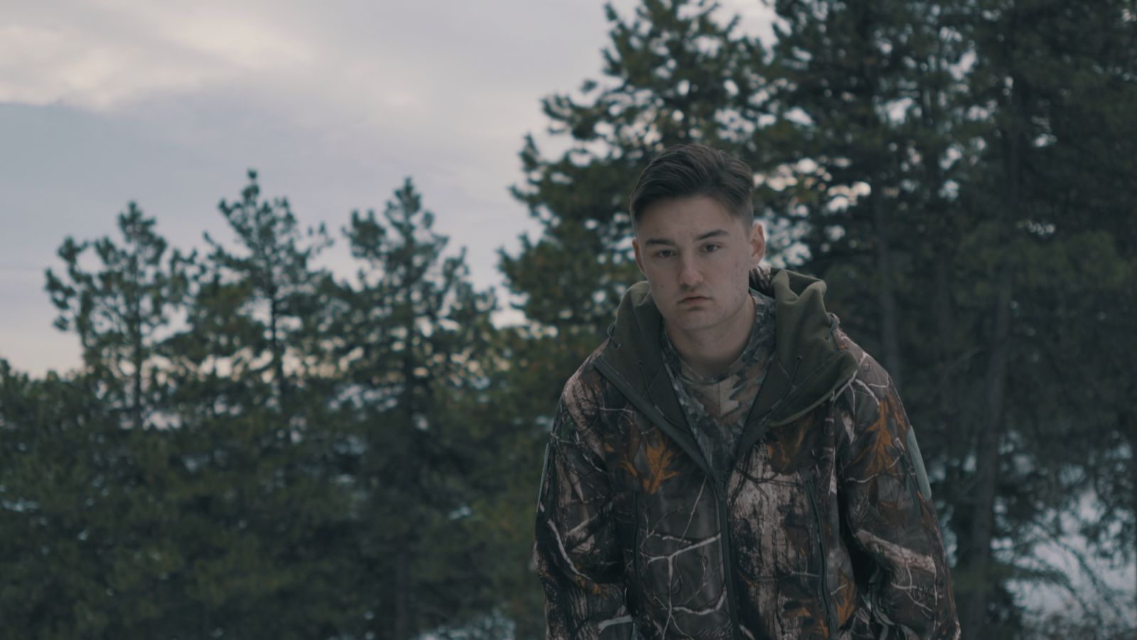 Angus wearing camouflage, trekking up a mountain in the snow while staring at the ground in the early evening