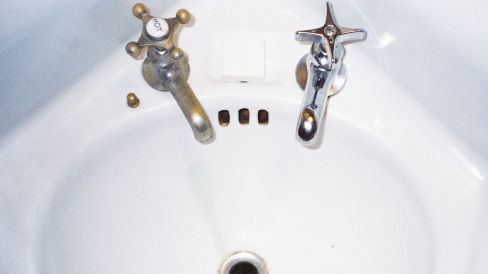 Film image of a porcelain sink from above. It has one gold faucet handle and one silver faucet handle. The entire image bleeds off the page, spanning across a 2-page spread.