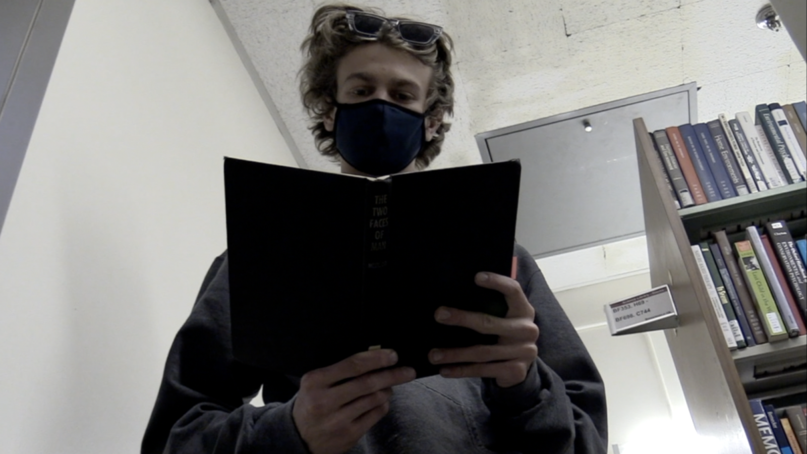 A corridor of books with strange titles like ‘Anatomy of Loneliness’ and ‘Humor and Laughter’. A lone man stands amongst them reading.