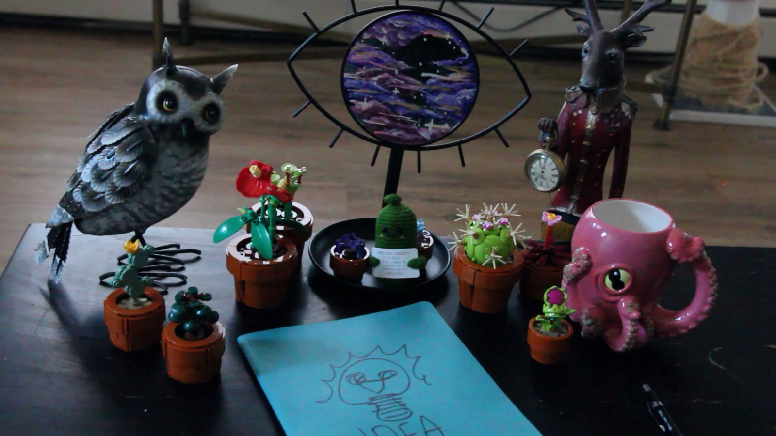 a shot of a bunch of objects surrounding an idea notebook. An owl, a metal eye, a deer statue, lego plants, and an octopus shaped mug