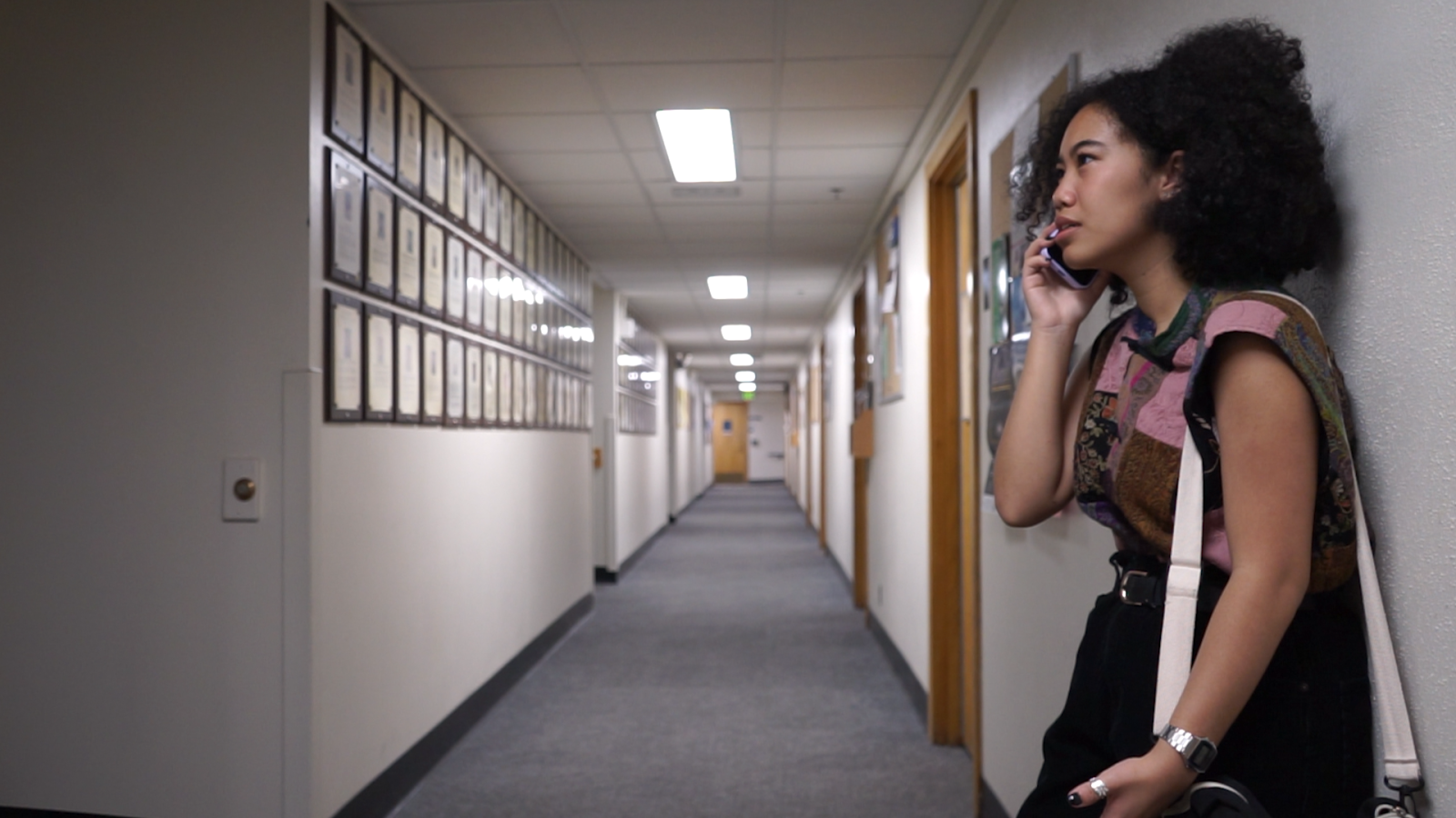 A woman leans against the wall of an empty hallway. She looks annoyed as she speaks to her father on the phone.