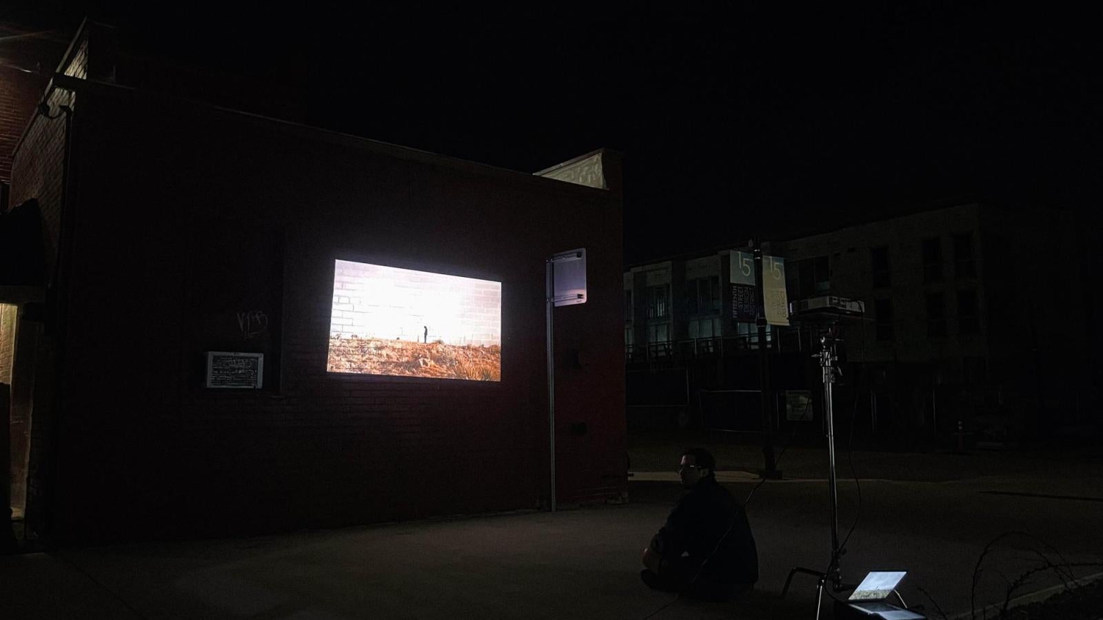 An image from the public installation of the project. A dark image with a brick building in the center with a bright image projected onto the center. To the right is a man seated crossed leg on the ground, his silhouette just visible. Behind him is a silver c stand with a projector on top of it attached to a laptop on the ground.