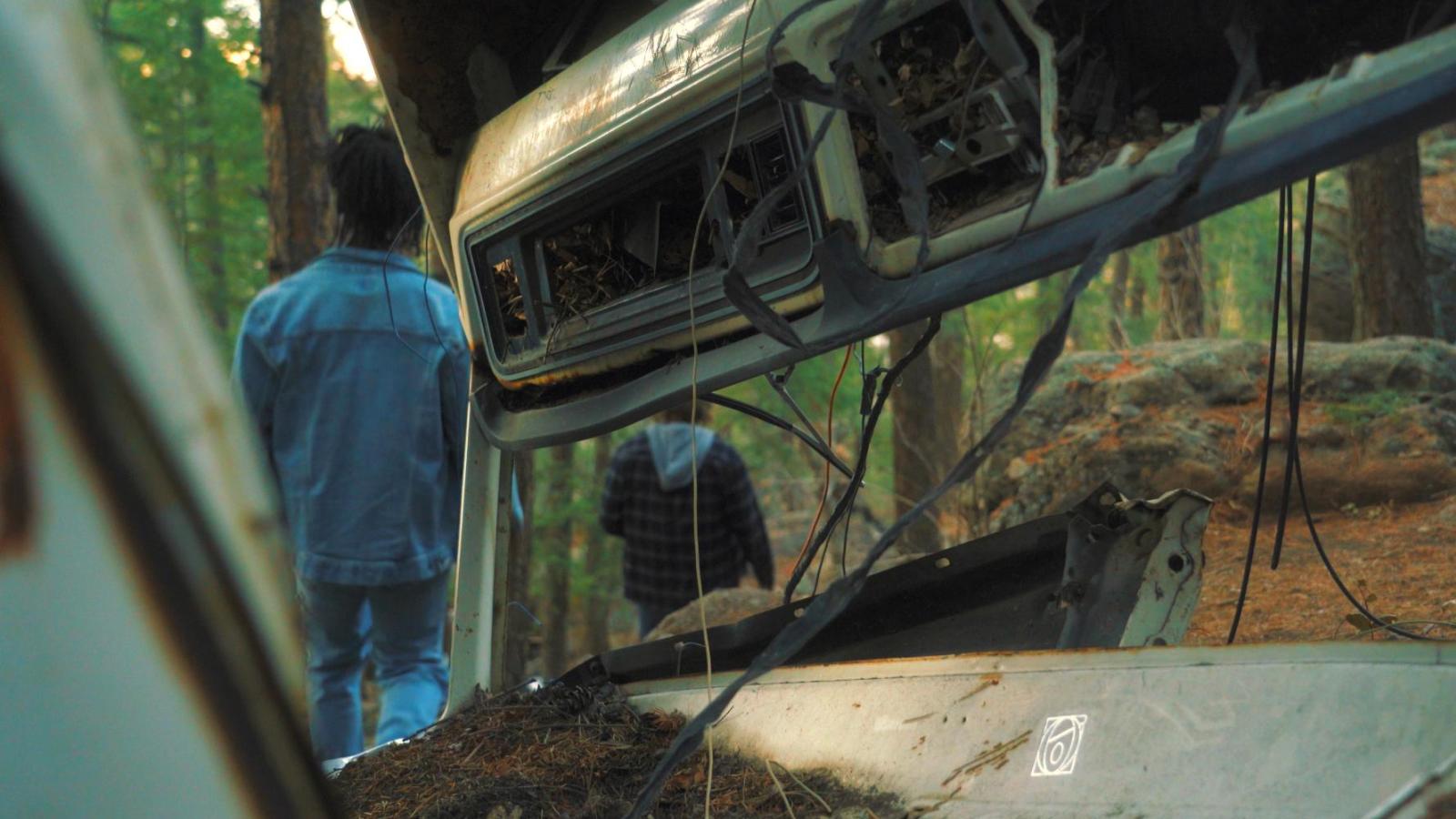 2 friends are seen walking past a car wrecked in the woods