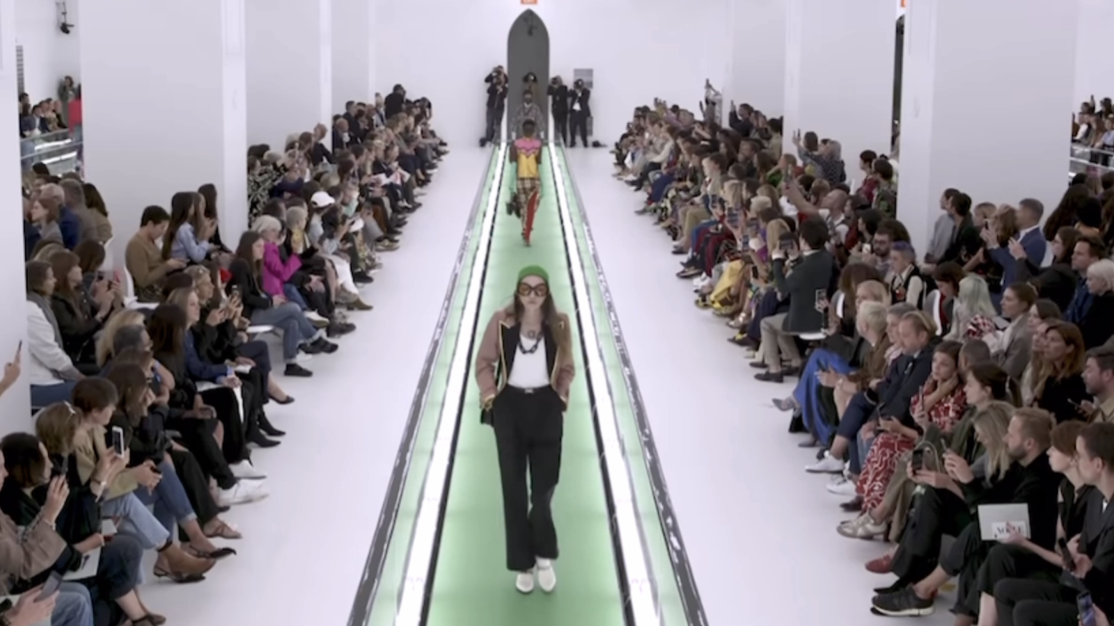 This photo is of a Gucci runway show. There are three models walking the runway and there are people lining the slim aisle.