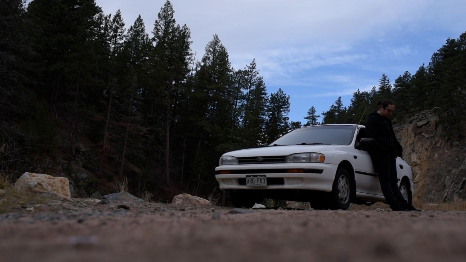 Person leaning against a car on a dirt road in the forest