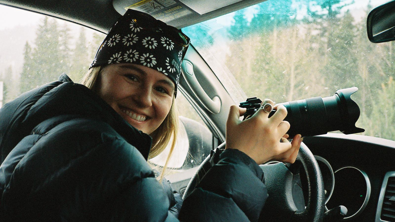 Person in drivers seat of car holding a camera and smiling