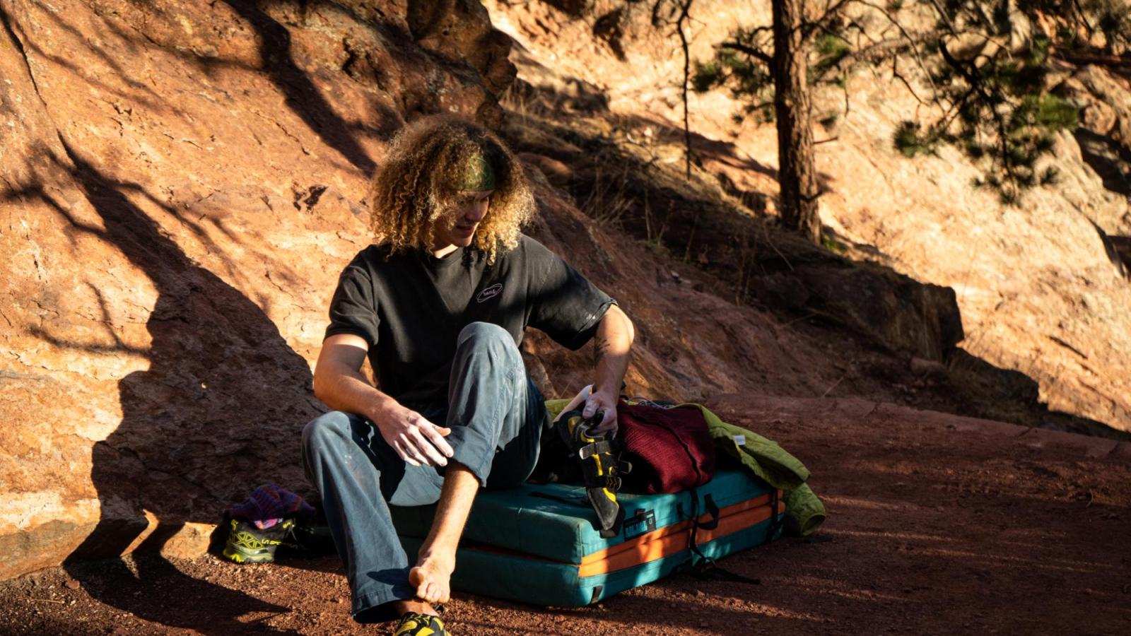 Shadows covering climber with curly hair and blue pants as he sits on pad to remove his shoes