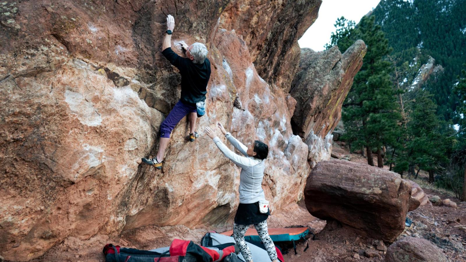 Elderly climber in purple pants reaching, while his wife in white spots him from behind