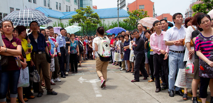 A group of people with umbrellas standing in line on a sunny day, waiting for their children to come out of an exam location of Chinese College Entrance Exam.