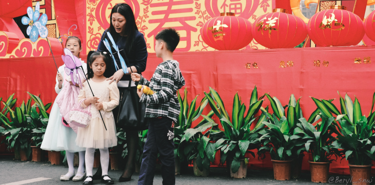 A woman and three children with toys stand in front of a red backdrop with lanterns and Chinese characters.