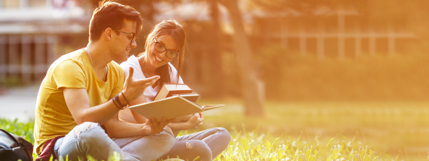 Two college students sit together in the grass studying textbooks happily.
