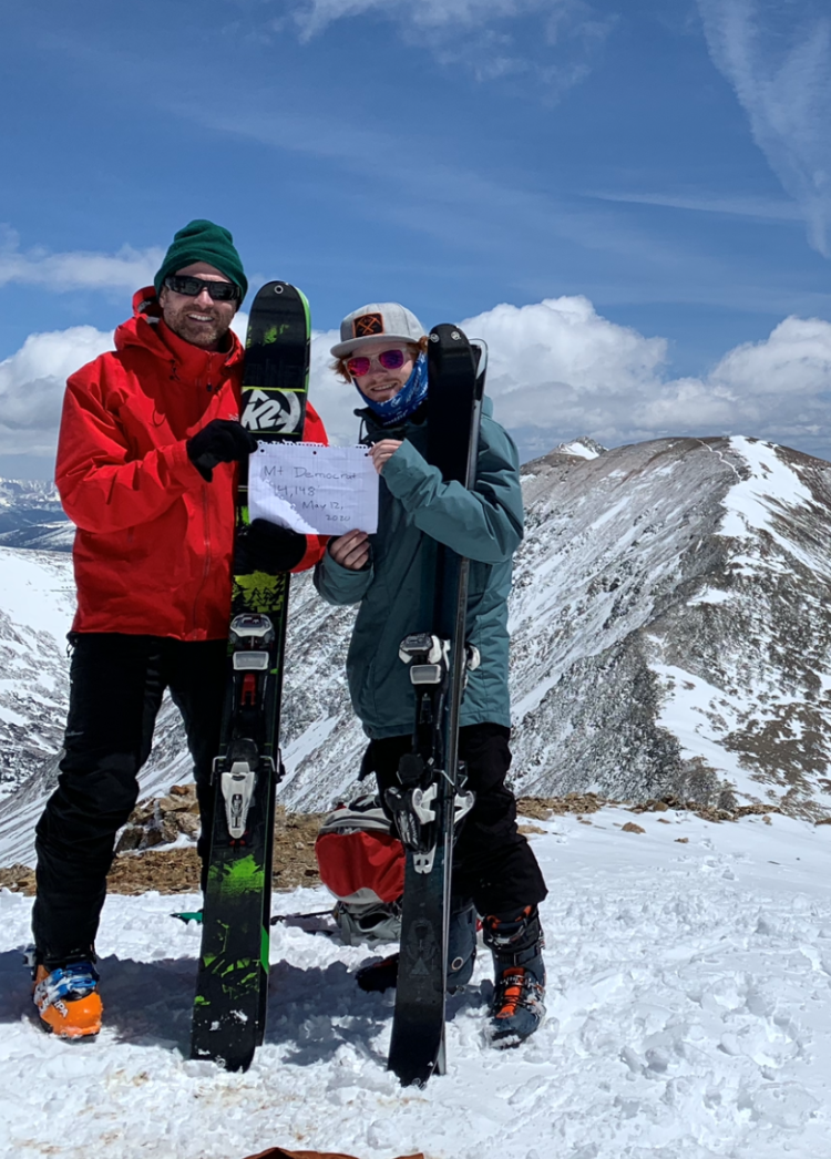 two men with skis and a sign at the top of a snowy mountain