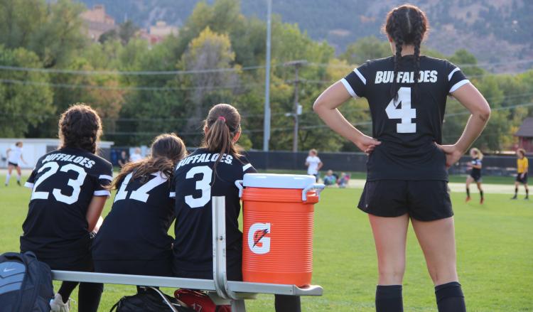 the back of four women's soccer players on the sideline during a game