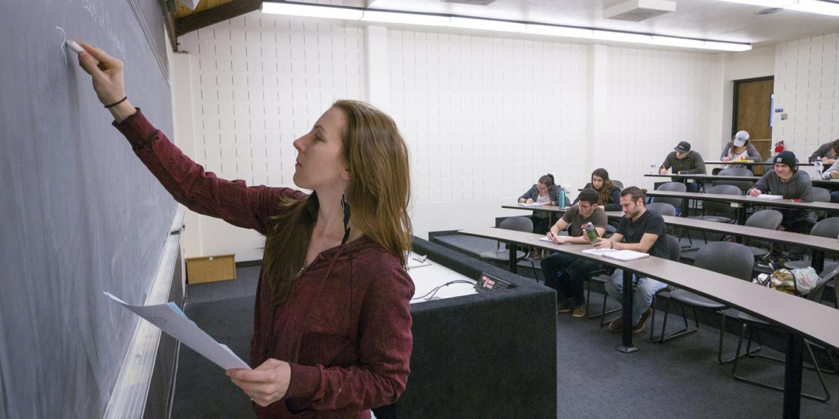 Photo of instructor writing on chalkboard with students at tables in background