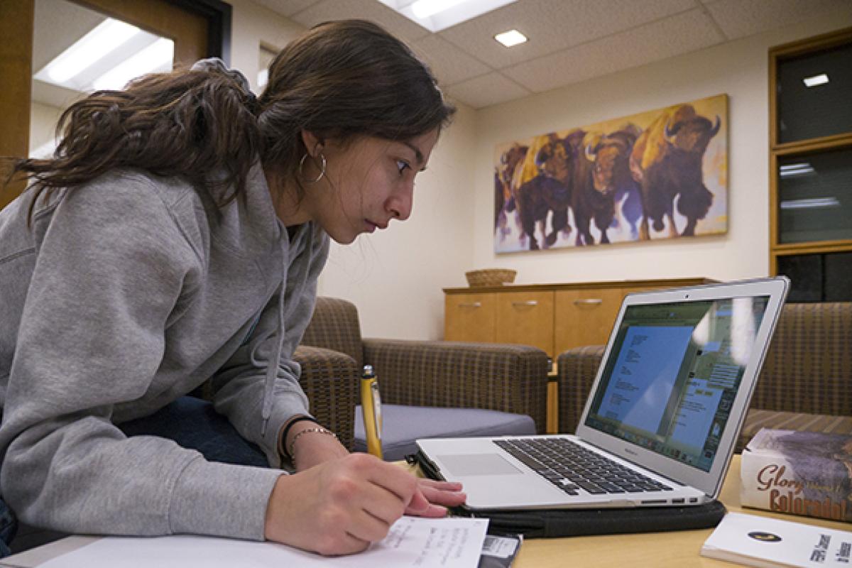 A student writes notes while consulting her laptop in the Office of the Registrar lobby.