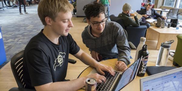 Two students work together on a laptop in a shared space.