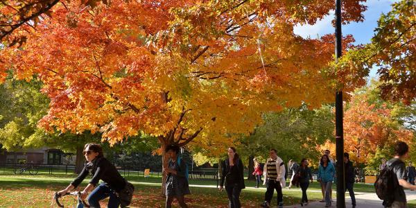 Students walk and bike across campus with fall foliage in the background.