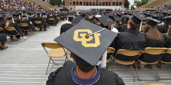 A view of the stage from the back of the student section at the spring commencement ceremony. In the foreground, a student is wearing a graduation cap with the CU logo on the top.