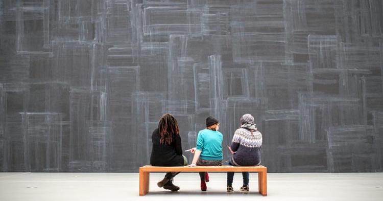 Three students sitting on a bench looking at a large wall with abstract line art.