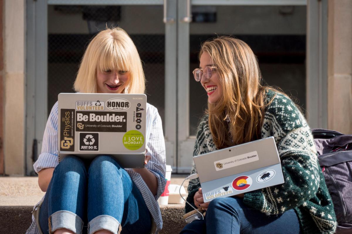 two students sitting on bench smiling, conversing, and looking at their laptops