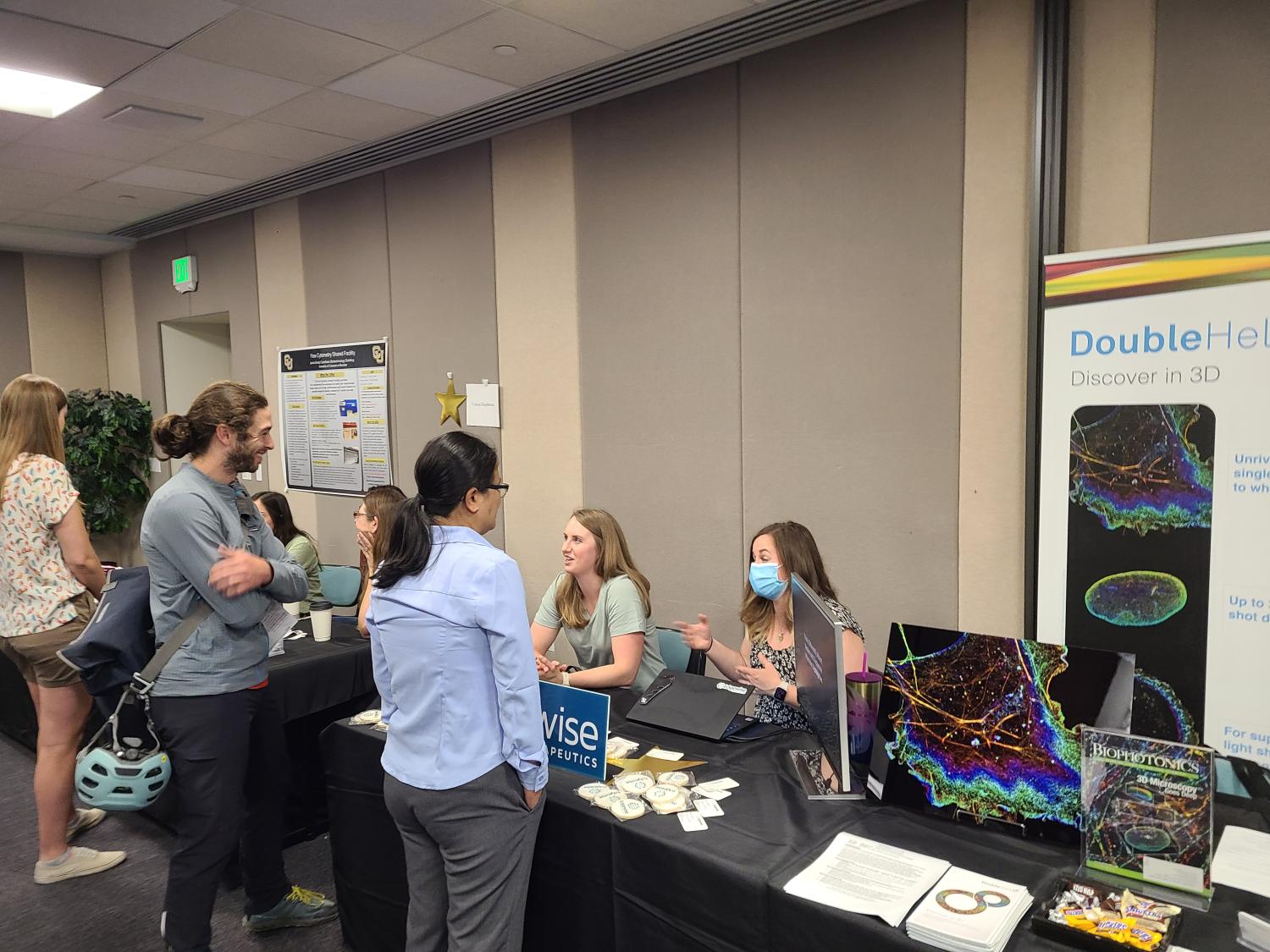 Image of two individuals standing while talking to two others behind a booth at the 2023 Core Facilities Open House.