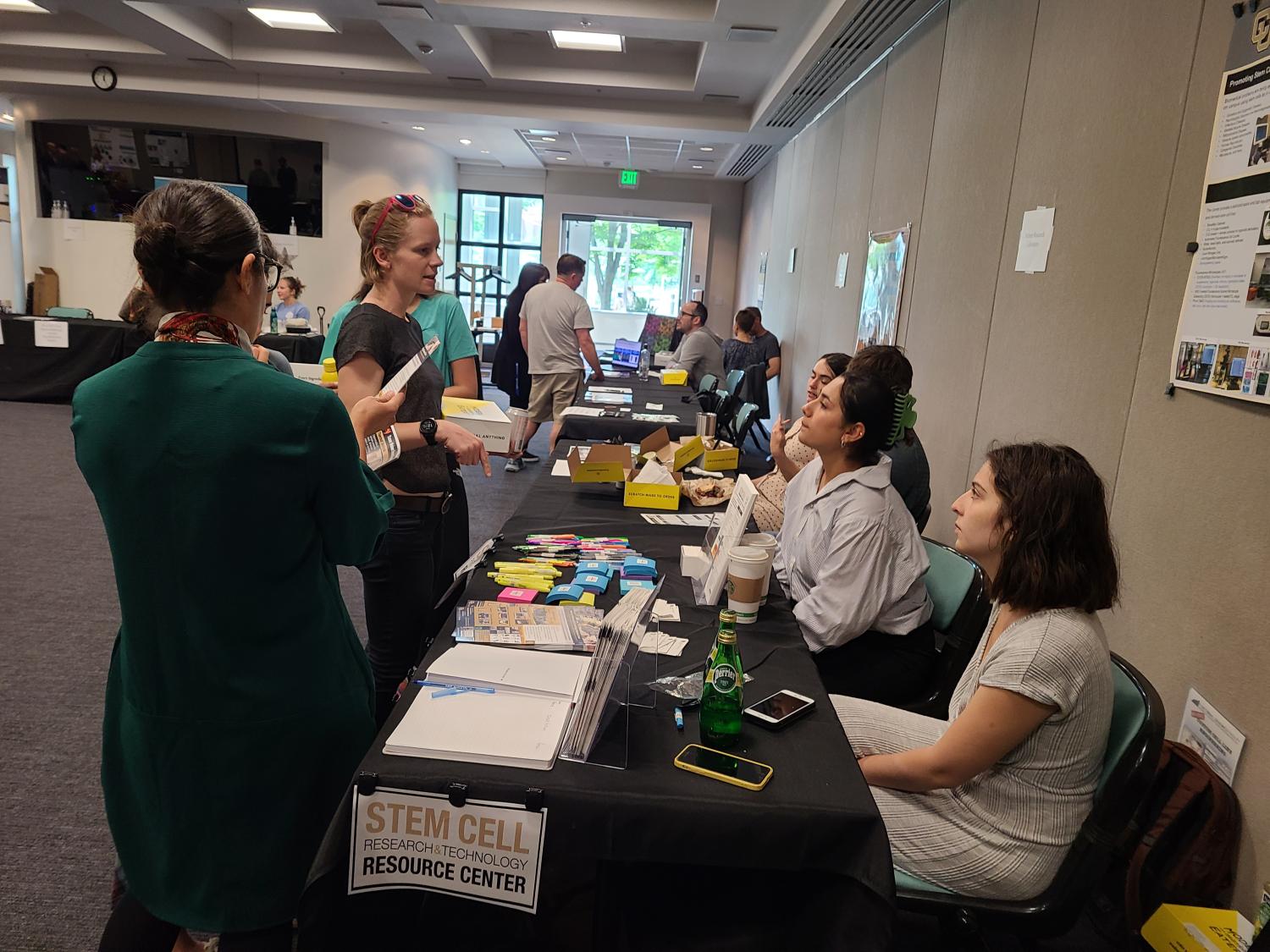 Image of two individuals standing while talking to two others behind a booth at the 2023 Core Facilities Open house.