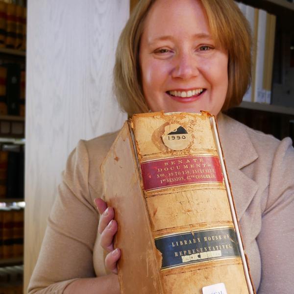 Kate Tallman is holding a thick volume of congressional records from the 1800s with bookshelves filled with books behind her in Norlin Library