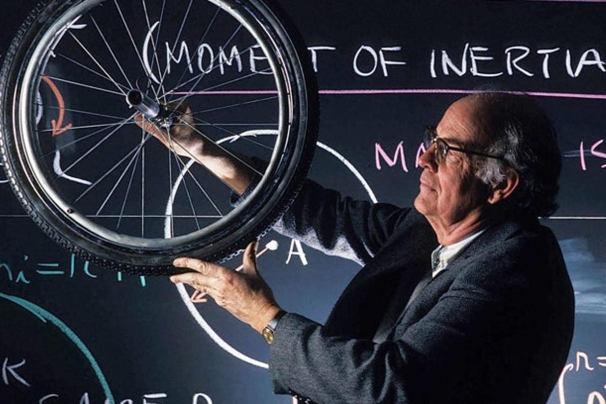 Physics professor holding a bicycle wheel in front of blackboard