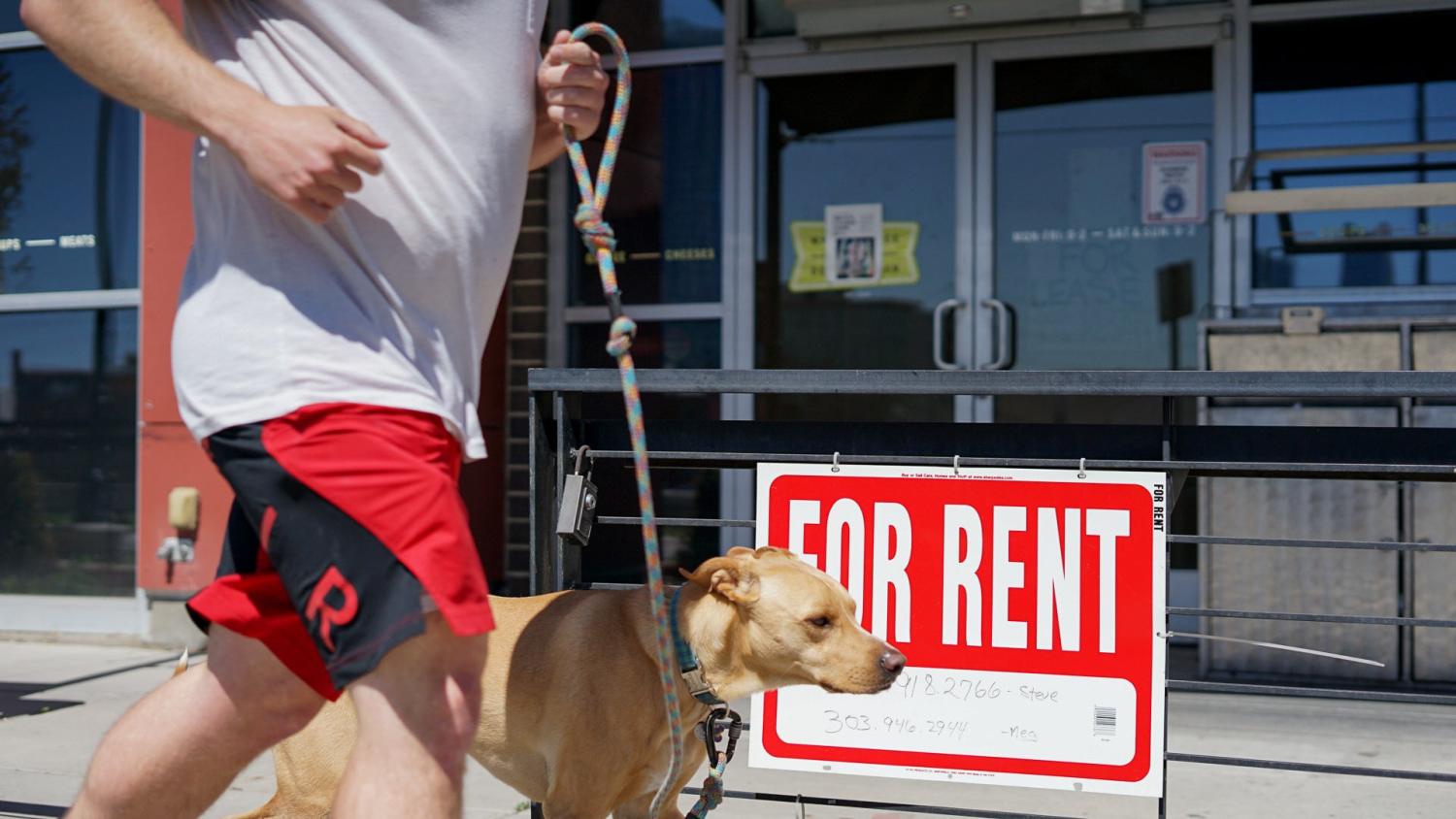A person and a dog run past a business displaying a 'For Rent' sign in Denver, Colorado.
