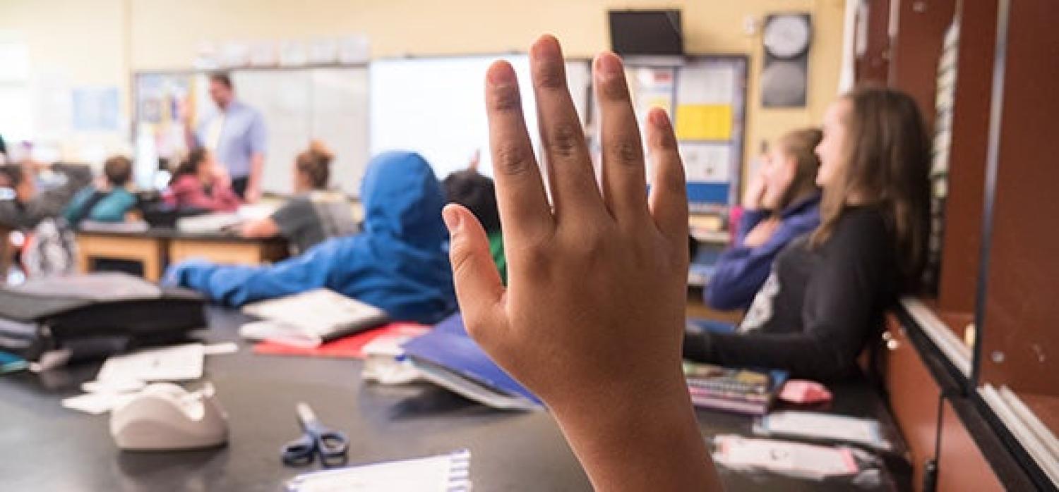 Elementary school students raising their hands with their teacher in the front of the class
