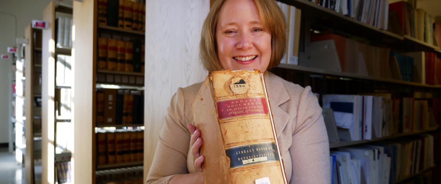 Kate Tallman is holding a thick volume of congressional records from the 1800s with bookshelves filled with books behind her in Norlin Library
