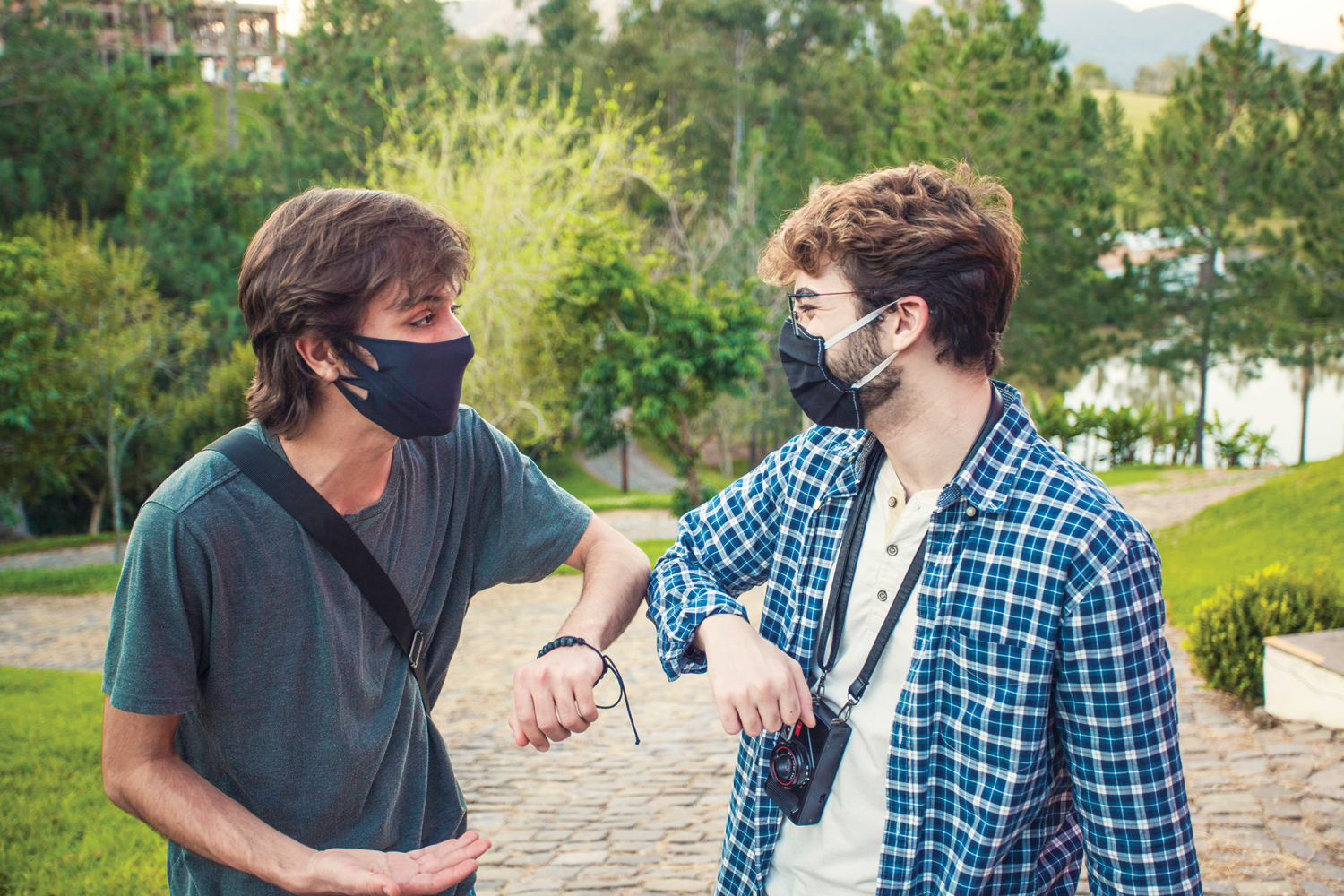 Two young men with masks bump elbows to greet each other in a park