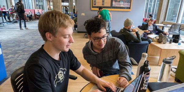 Two students work together on a laptop in a shared space.