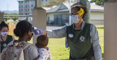 A woman conducts a temperature check of a mother and her baby while wearing a mask.