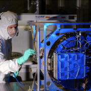 Engineer in protective gear inspects a metal space instrument with wires coming out of its back