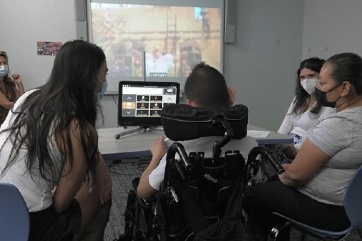 An adolescent in a wheelchair using eye gaze to access his AAC system, while his mother and two graduate clinicians show him comments on the AAC system about the movie Encanto that’s playing on the screen.