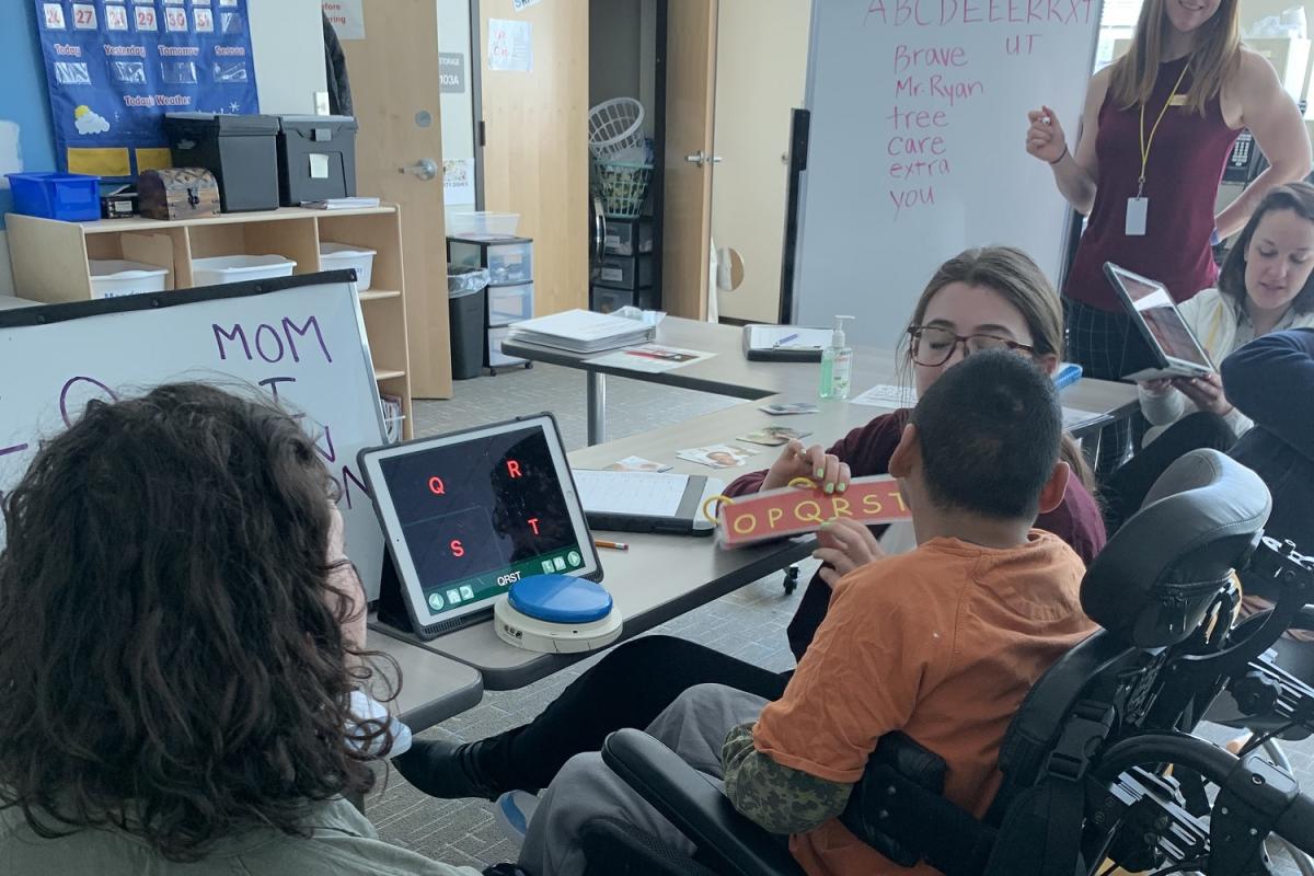 A graduate student clinician is helping an adolescent write as he selects letters on an alphabet flip chart that she is presenting to him.
