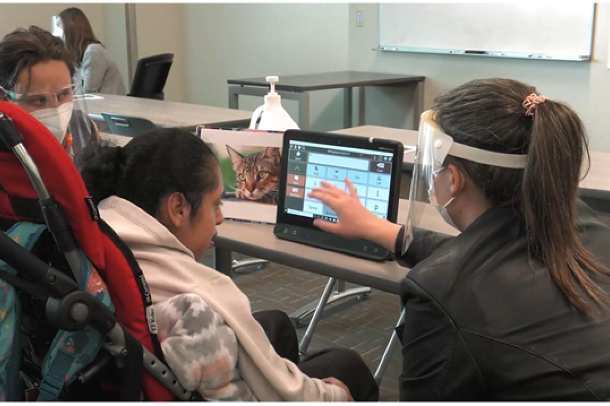 Two graduate students are helping an adolescent use the keyboard on her AAC system to write about cats.