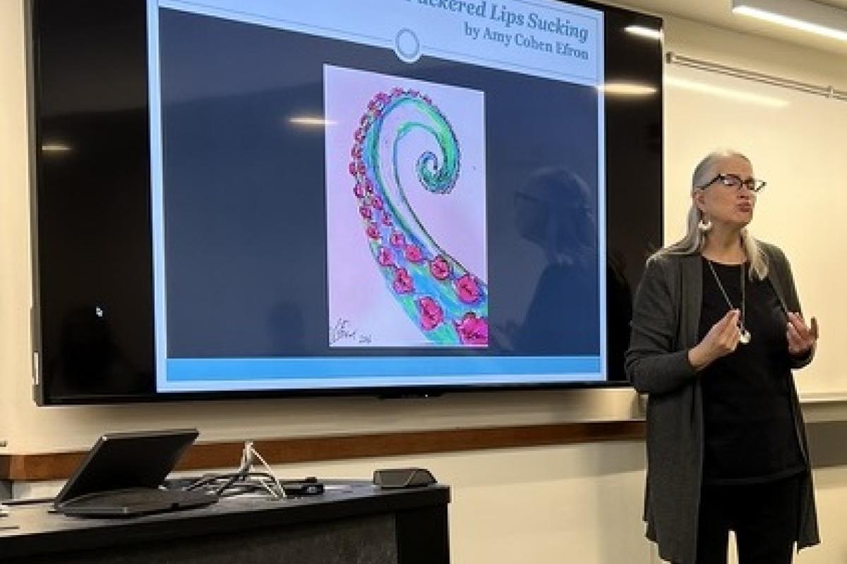 ASL Instructor teaching ASL 1 to students showing a slide in the back with “Puckered Lips Sucking” by Amy Cohen Efron and the instructor is showing "puckered" lips with her facial expression.