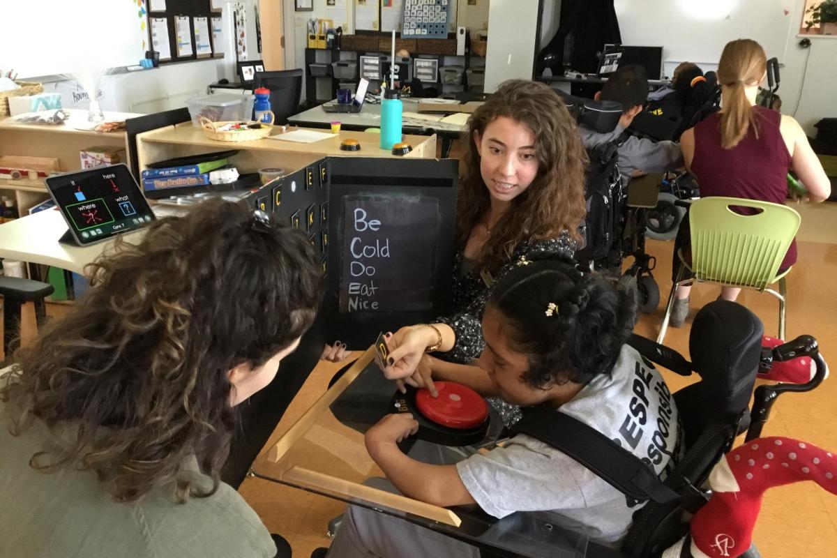 Two graduate student clinicians helping an adolescent with writing by presenting the full alphabet to her on a large trifold and scanning the letters verbally while she selects letters she wants by pushing a single message communicator placed in front of her, "That's the one I want."