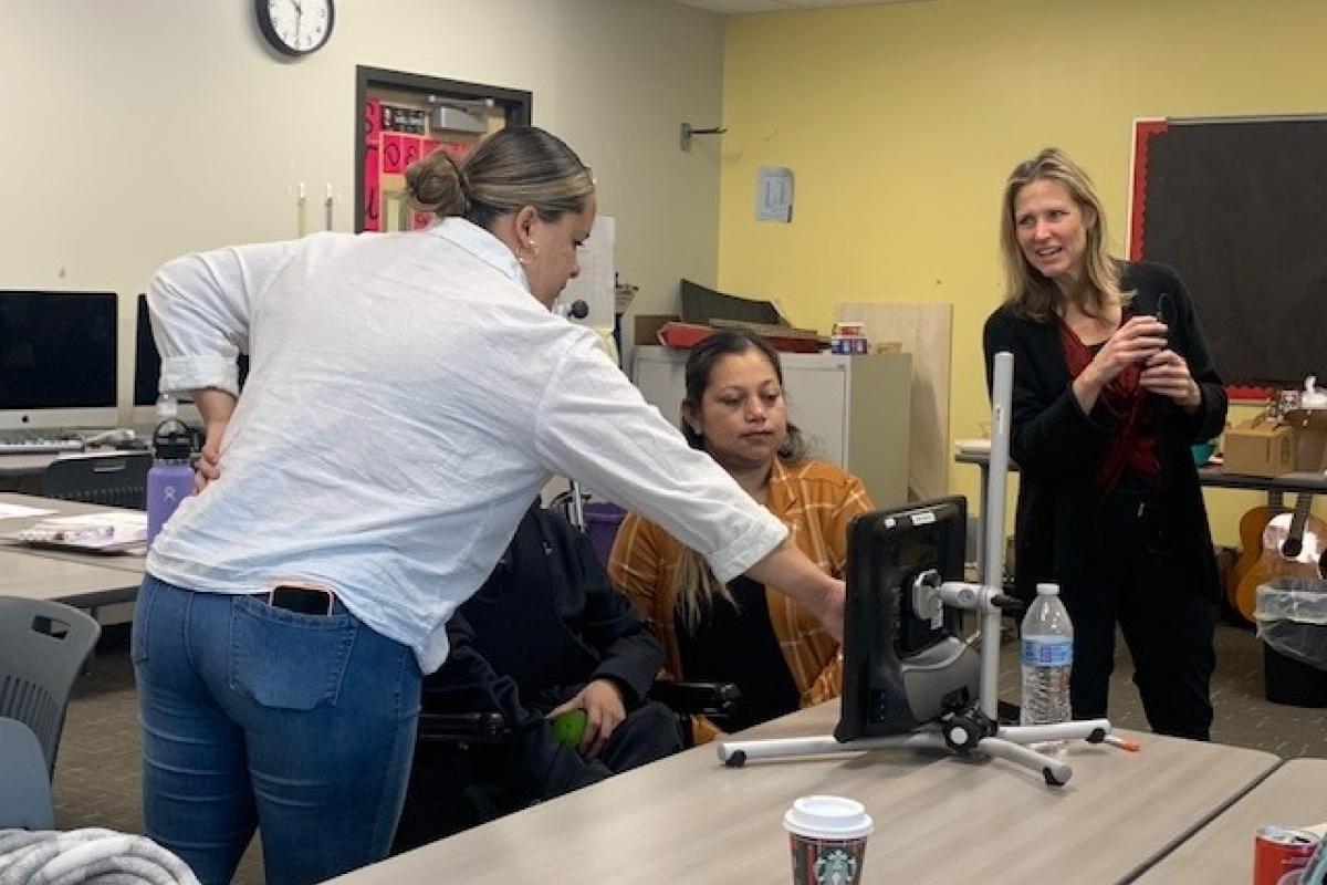 A clinical faculty member coaching a graduate student clinician who is showing the mother of an adolescent and the adolescent where vocabulary is on his AAC device.